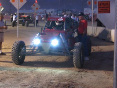 1990 Ford Bronco - red off-road race buggy with headlights lit idles on dirt at night, its helmeted driver aboard and a man standing beside it.