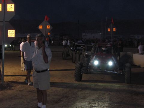 1990 Ford Bronco - off-road race buggy advances with bright headlights on a dirt course at dusk past spectators and lit arrow course markers.