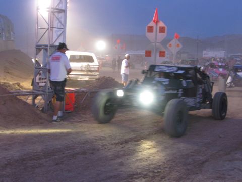 1990 Ford Bronco - black off-road race buggy drives with twin headlights past a dirt berm at dusk while crew and spectators look on.