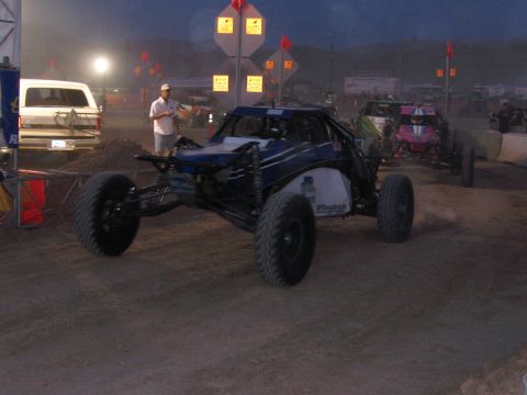 1990 Ford Bronco - blue-and-white tube-frame off-road race buggy with knobby tires moves along a dirt course at dusk near a marshal.
