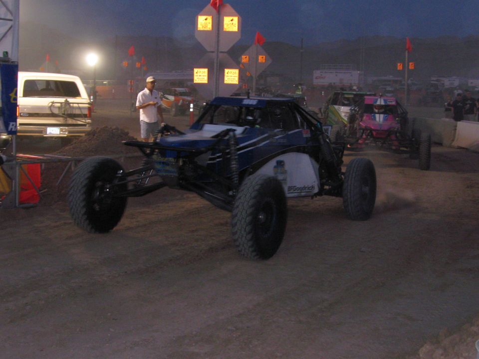 Blue and white off-road buggy with knobby tires on a dirt course at dusk