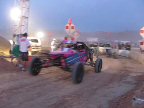 1990 Ford Bronco - pink-and-blue tube-frame off-road race buggy with knobby tires moves along a dusty course at dusk past course markers.
