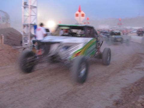 1990 Ford Bronco - white-and-green off-road race buggy kicks up dust as it powers along a dirt course at dusk near other buggies.