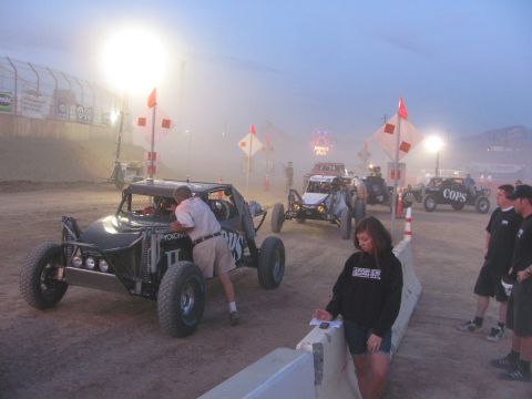 1990 Ford Bronco - black off-road race buggy and others sit on a dusty staging course at dusk as spectators and crew stand nearby.