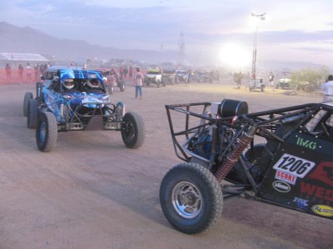 1990 Ford Bronco - blue off-road buggy and a numbered tube-frame race car sit on a dusty desert lot at dusk with mountains and bright lights behind.
