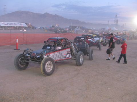 1990 Ford Bronco - Checkered-paint off-road race buggies line up on a dusty desert lot at dusk beside orange safety fencing and crew.