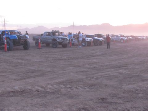 1990 Ford Bronco - long row of off-road race trucks and buggies stages on a dusty desert lot at dawn with mountains and spectators behind.