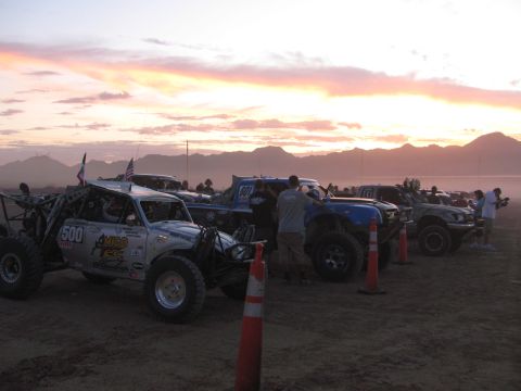 1990 Ford Bronco - row of off-road race trucks with flags lines up on a dusty desert lot under a vivid pink-and-orange sunset sky.