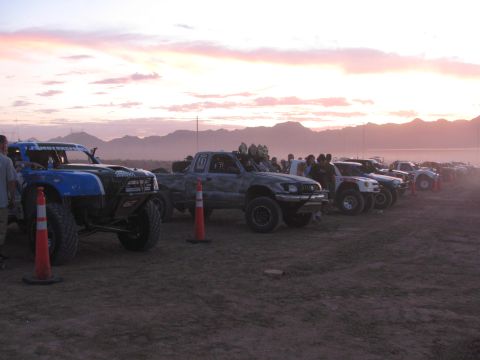 1990 Ford Bronco - line of off-road race trucks stages on a dusty desert lot at sunset, traffic cones in front and mountains behind.