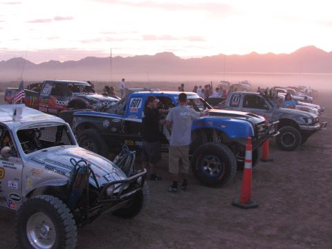 1990 Ford Bronco - Off-road race trucks and buggies with sponsor decals sit on a dusty desert lot at sunset as crew work between them.