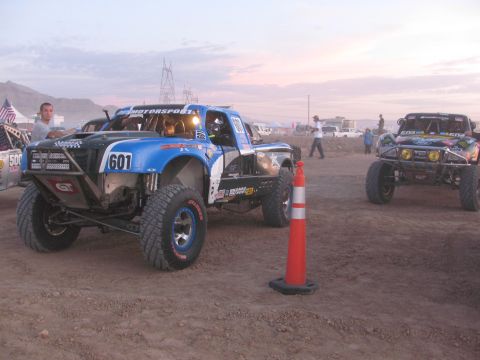 1990 Ford Bronco - blue and white tube-chassis off-road race truck marked 601 sits on a dusty staging lot at dusk with another race truck nearby.