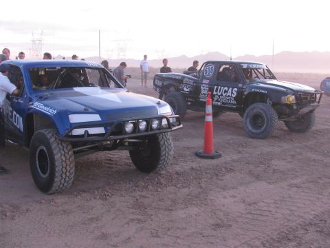 1990 Ford Bronco - blue off-road race truck parked next to a darker race truck marked 606 with Lucas sponsorship on a dusty staging area.