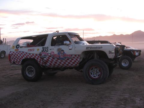 1990 Ford Bronco - white off-road race SUV with red checkerboard graphics, numbered 303 and labeled Big n Boy, staged on a dusty lot at dusk.