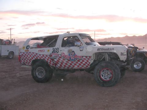 1990 Ford Bronco - white off-road race SUV with red checkered graphics numbered 303 sits on a dusty lot with a utility service truck behind it.