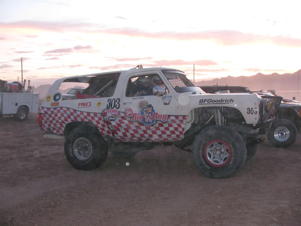 White and red checkered race SUV number 303 staged on dirt with a service truck behind