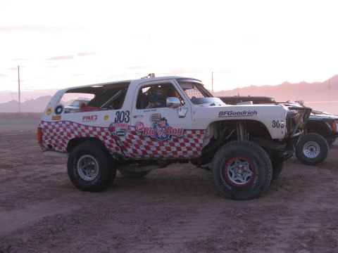 1990 Ford Bronco - Side view of a white off-road race SUV marked 303 with red checkerboard and Big n Boy graphics on a dusty staging lot at dusk.