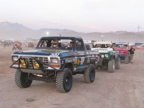 1990 Ford Bronco - row of off-road race vehicles staged in the desert at dusk, led by a black race pickup marked 300.