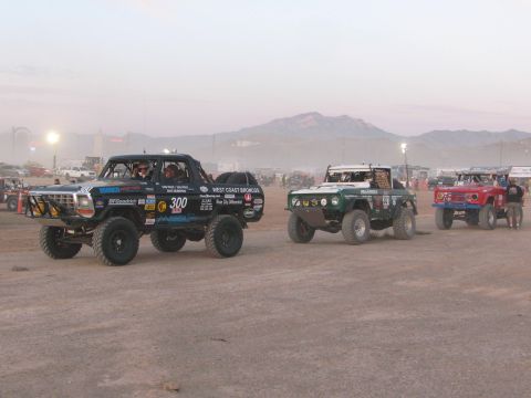 1990 Ford Bronco - black off-road race pickup numbered 300 lines up with other race vehicles on a dusty desert flat at dusk.