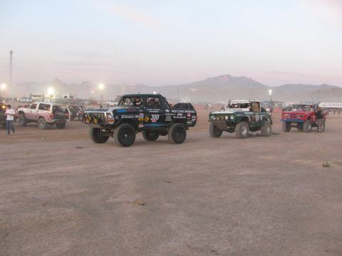 1990 Ford Bronco - Several off-road race vehicles, including a black pickup marked 300, line up on a dusty desert lot at dusk with mountains behind.