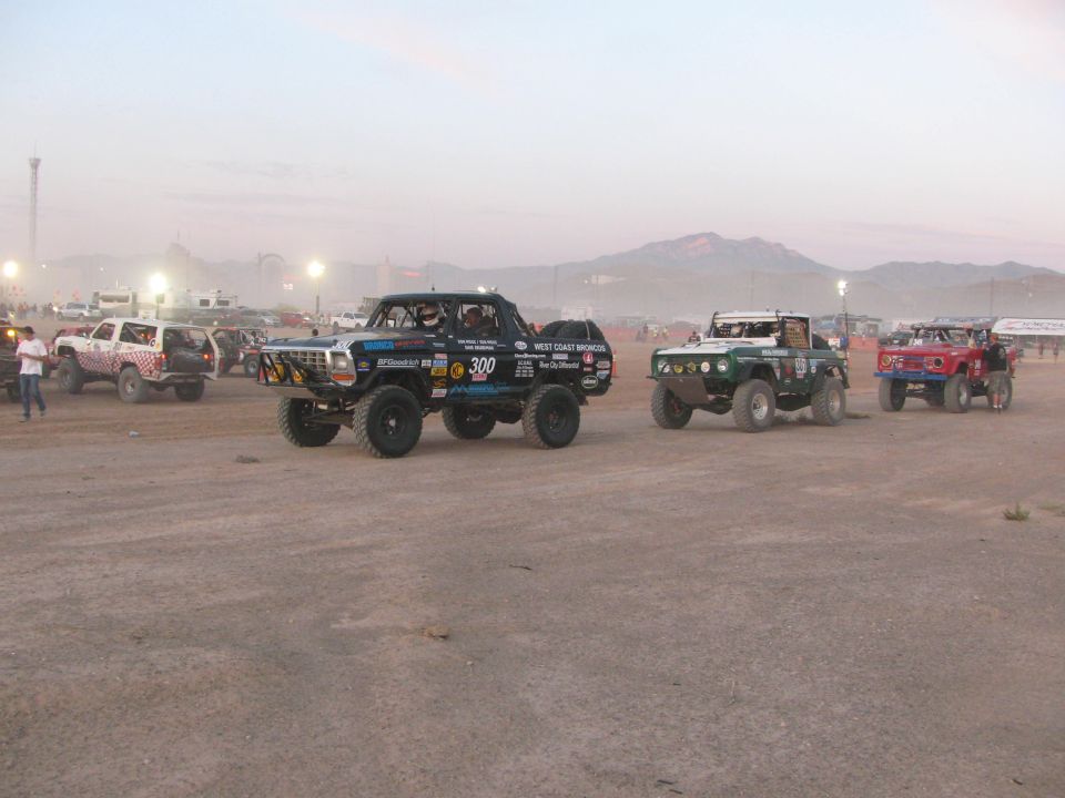 Several off-road race vehicles staged in a row on dusty desert ground at dusk
