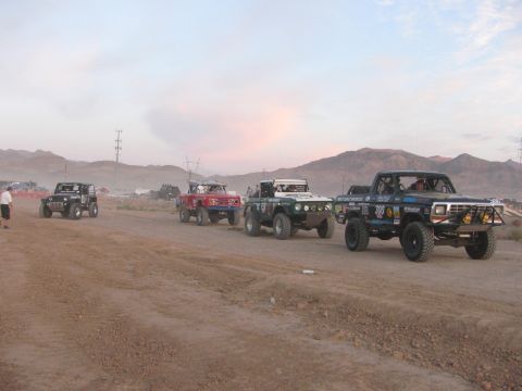 1990 Ford Bronco - row of off-road race trucks parked along a dirt road at dusk, the nearest a black pickup marked 300.