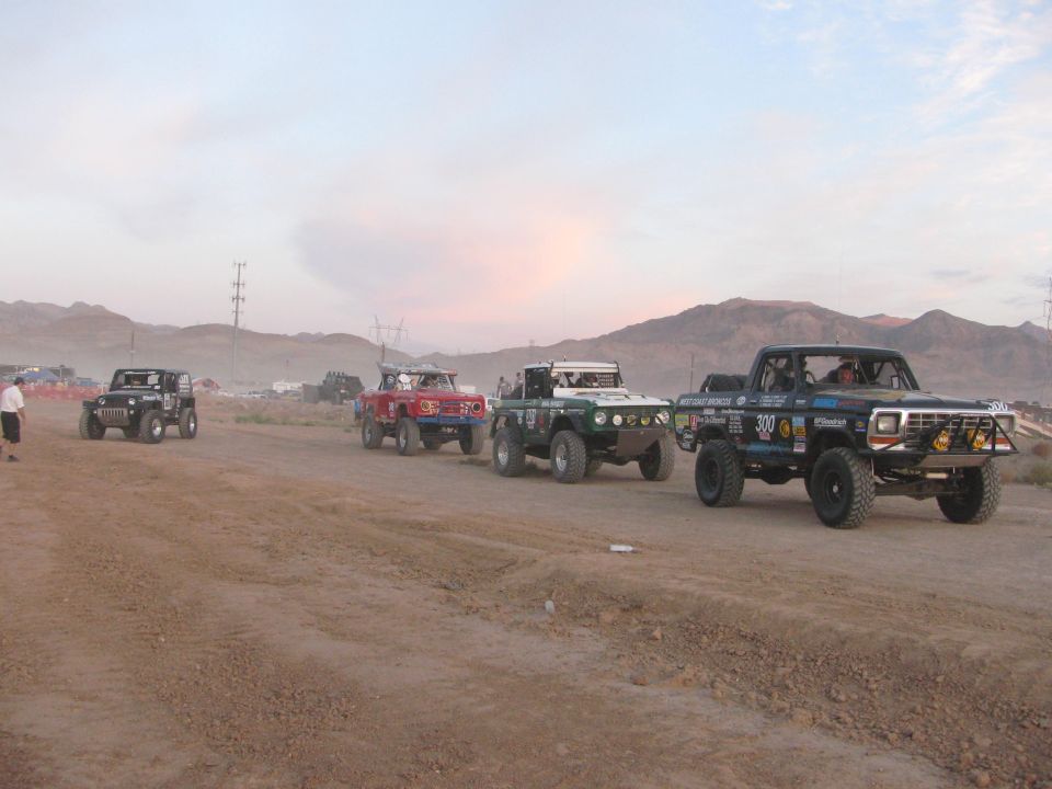 Row of off-road race trucks parked along a dirt road at dusk with mountains behind