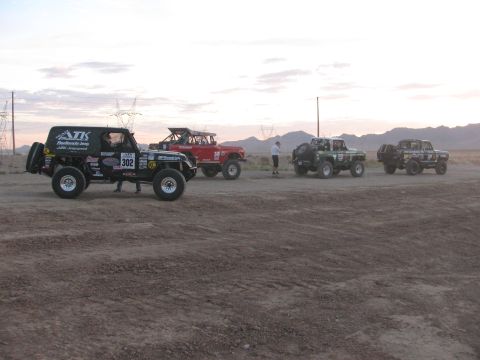 1990 Ford Bronco - black off-road race Jeep numbered 302 leads a line of staged race vehicles across a dusty desert flat at dusk.