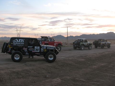1990 Ford Bronco - black off-road race Jeep marked 302 stages alongside other race vehicles on a dusty desert lot under a sunset sky.