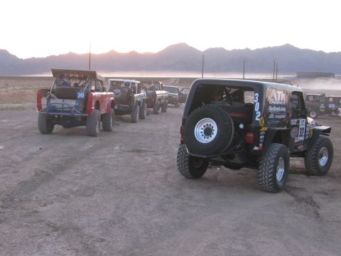 1990 Ford Bronco - Rear view of a black off-road race Jeep marked 302 with a spare tire, queued behind other race vehicles at dawn.