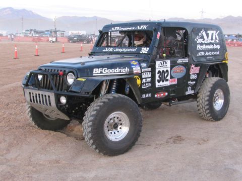 1990 Ford Bronco - black off-road race Jeep numbered 302 with a helmeted driver and ATK Jeepspeed graphics sits on a dusty staging lot.