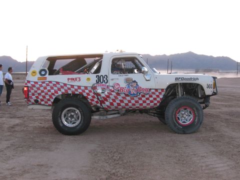 1990 Ford Bronco - Side view of a white off-road race SUV marked 303 with red checkerboard graphics parked on a dusty lot at dusk.