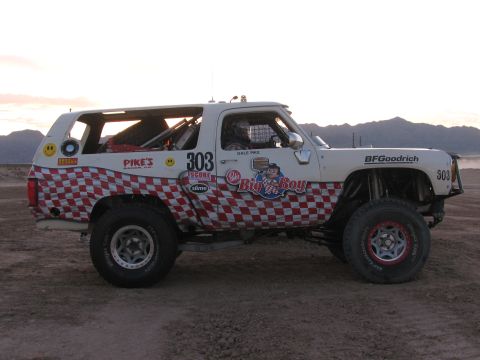 1990 Ford Bronco - white off-road race SUV numbered 303 with red checkered and Big n Boy graphics staged on a desert lot at dusk.