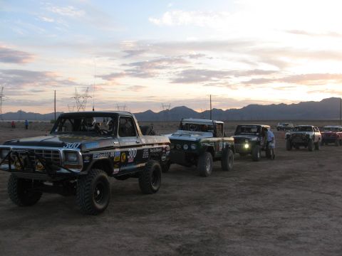 1990 Ford Bronco - line of off-road race trucks led by a black pickup marked 300 staged in the desert under a sunset sky.