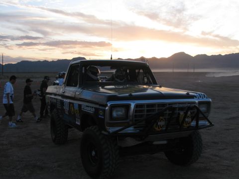 1990 Ford Bronco - black off-road race pickup marked 300 sits on a desert flat at sunset as several people stand alongside it.