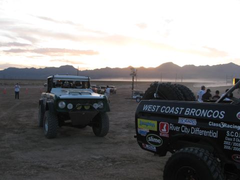 1990 Ford Bronco - green off-road race vehicle stages beside the rear of a West Coast Broncos race truck on a dusty desert flat at sunset.