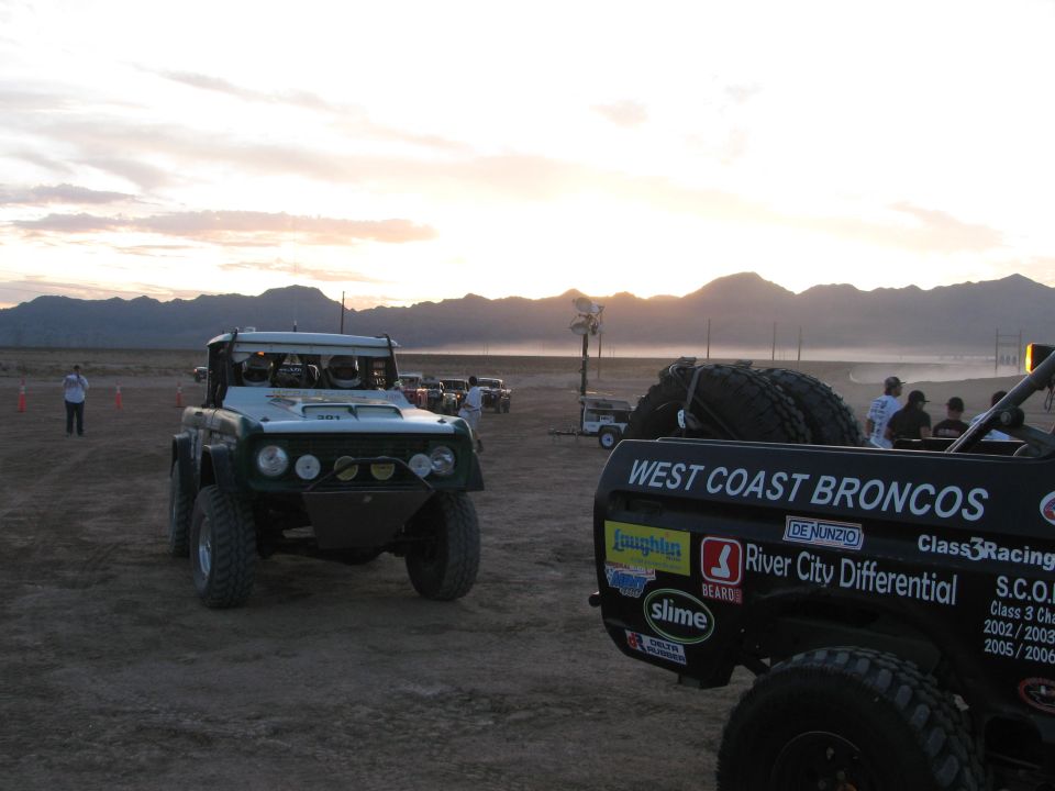 Green off-road race vehicle and a West Coast Broncos truck rear staged at sunset