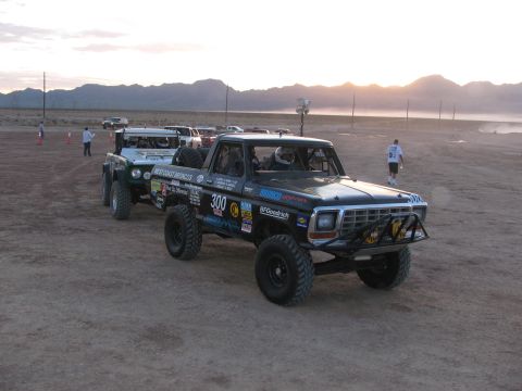 1990 Ford Bronco - black off-road race pickup marked 300 stages near a green West Coast Broncos truck on a hazy desert flat at sunset.
