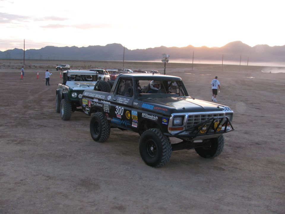 Black off-road race pickup number 300 and a green truck staged in the dusty desert