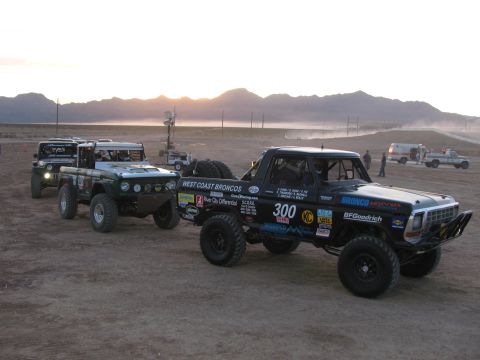 1990 Ford Bronco - black West Coast Broncos race truck marked 300 stages with green off-road trucks on a hazy desert flat at sunset.