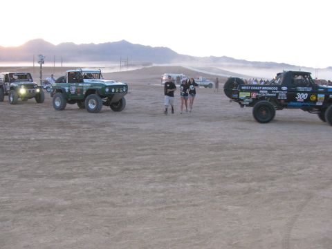 1990 Ford Bronco - Two people walk among staged off-road race vehicles, including a green truck and West Coast Broncos pickup 300, on a hazy desert flat.