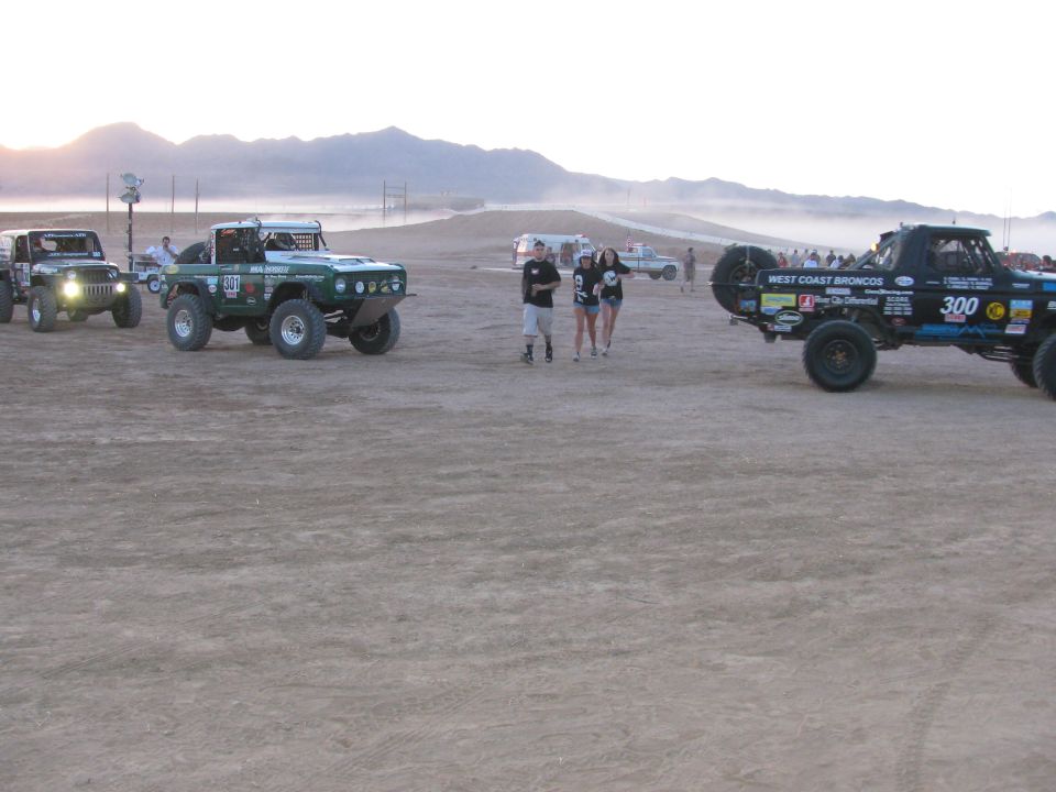 People walking among staged off-road race vehicles on a dusty desert flat at sunset