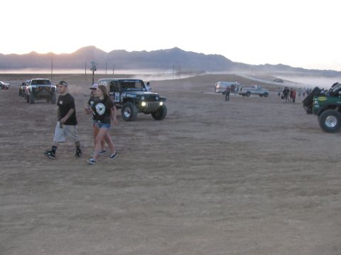 1990 Ford Bronco - Two people walk past a white off-road race Jeep with other race vehicles scattered across a hazy desert flat at sunset.