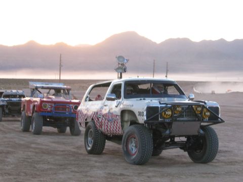 1990 Ford Bronco - white off-road race SUV marked 303 with checkered graphics stages near a red vehicle on a hazy desert flat at dawn.
