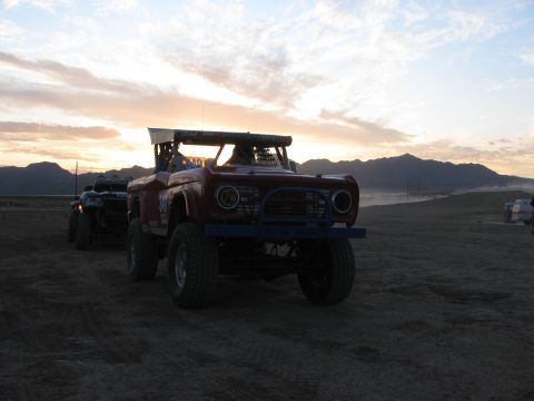 1990 Ford Bronco - red early-Bronco-style off-road race truck idles on a desert course at dusk with mountains and sunset clouds behind.