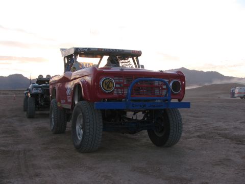 1990 Ford Bronco - red early-Bronco off-road racer with a blue tube front bumper and 'Ford' lettering sits at a dusty staging area at sunset.
