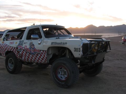 1990 Ford Bronco - white off-road race truck numbered 303 with red checkered graphics and BFGoodrich decals sits on a dusty course at dusk.