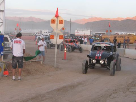 1990 Ford Bronco - number 551 off-road Baja-style racer waits at a dusty start area as a SCORE official holds a green flag amid spectators.