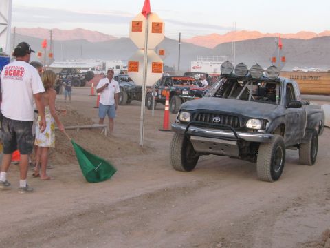 1990 Ford Bronco - gray Toyota Tacoma prerunner with a roof-mounted light bar rolls past officials and spectators at a dusty desert staging area.
