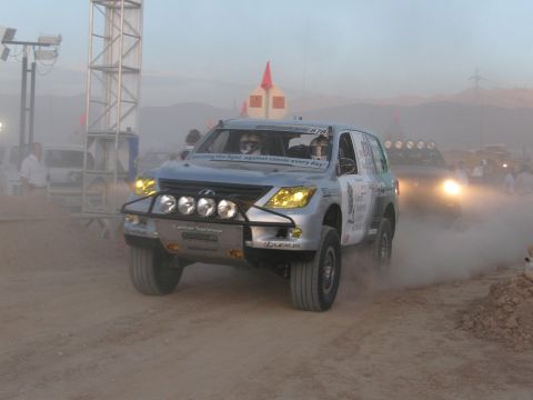 1990 Ford Bronco - silver Lexus off-road race SUV with cancer-charity decals kicks up dust mid-course at dusk under stadium lighting.