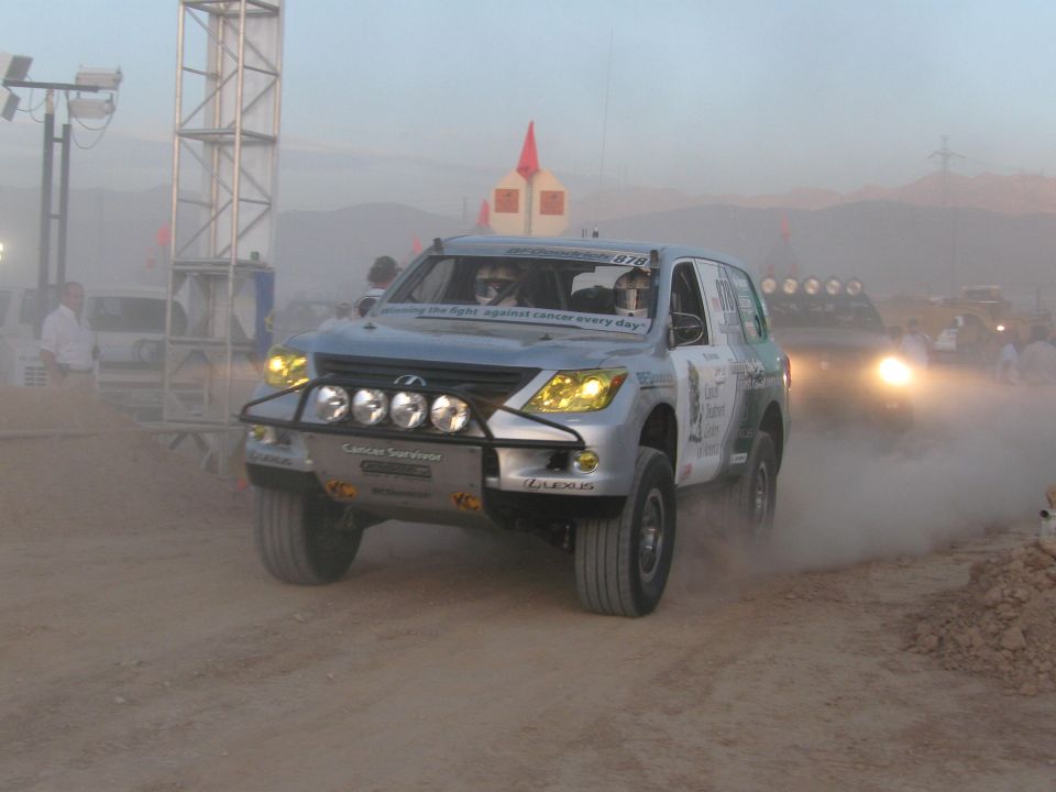 Silver Lexus LX off-road race SUV jumping through dust with headlights on